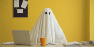 Person wearing ghost costume sitting at office desk with laptop and coffee mug, representing IT nightmares at work